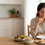 Adult still feeling hungry after a meal beside balanced foods containing protein, fiber-rich carbohydrates, vegetables, fruit, and healthy fats